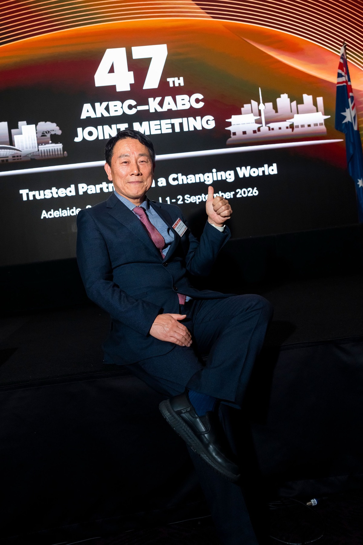A delegate giving a thumbs up in front of the 47th AKBC-KABC Joint Meeting stage backdrop, Adelaide